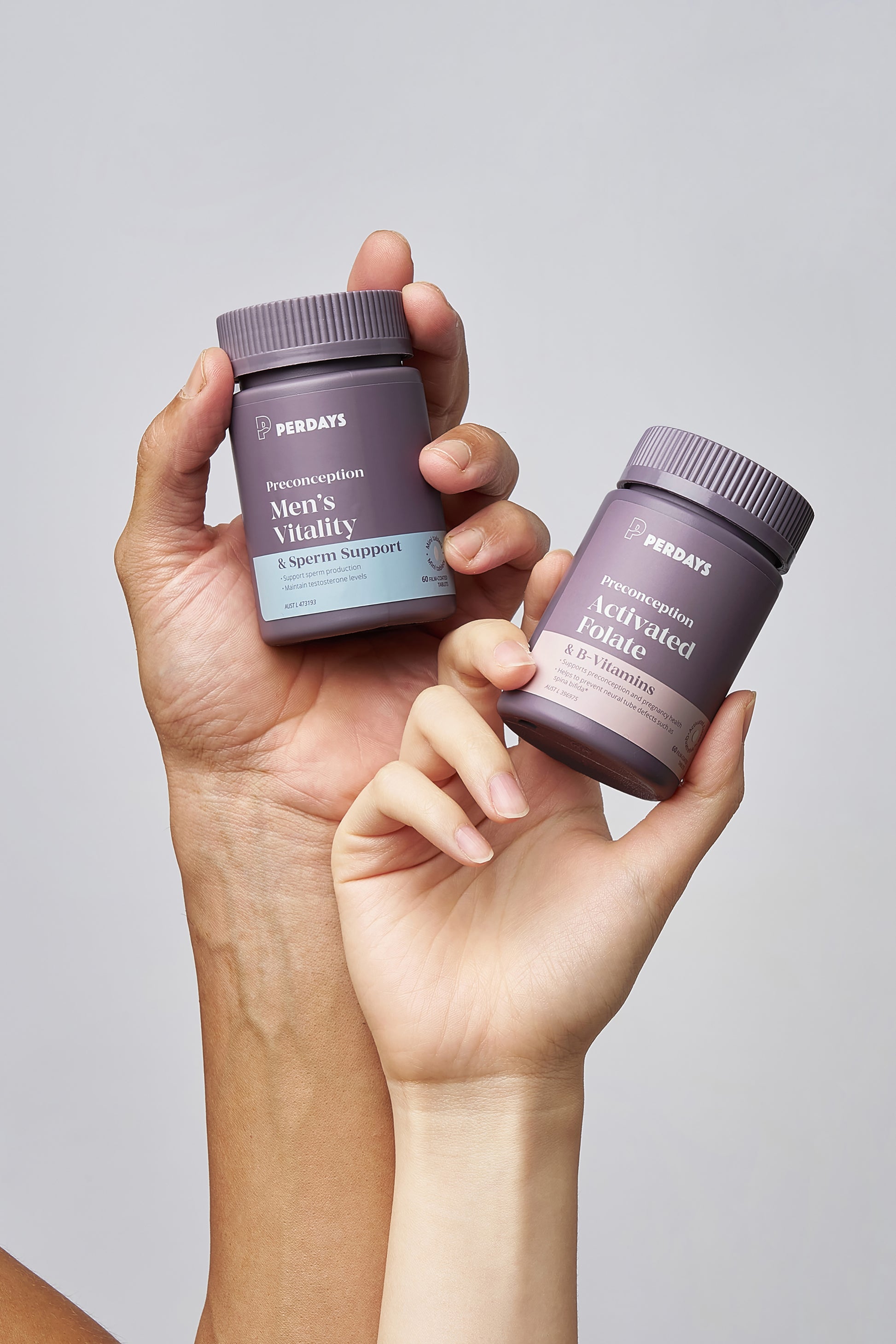 Two hands holding two preconception purple supplement bottles labeled 'Perdays' on a light gray background.